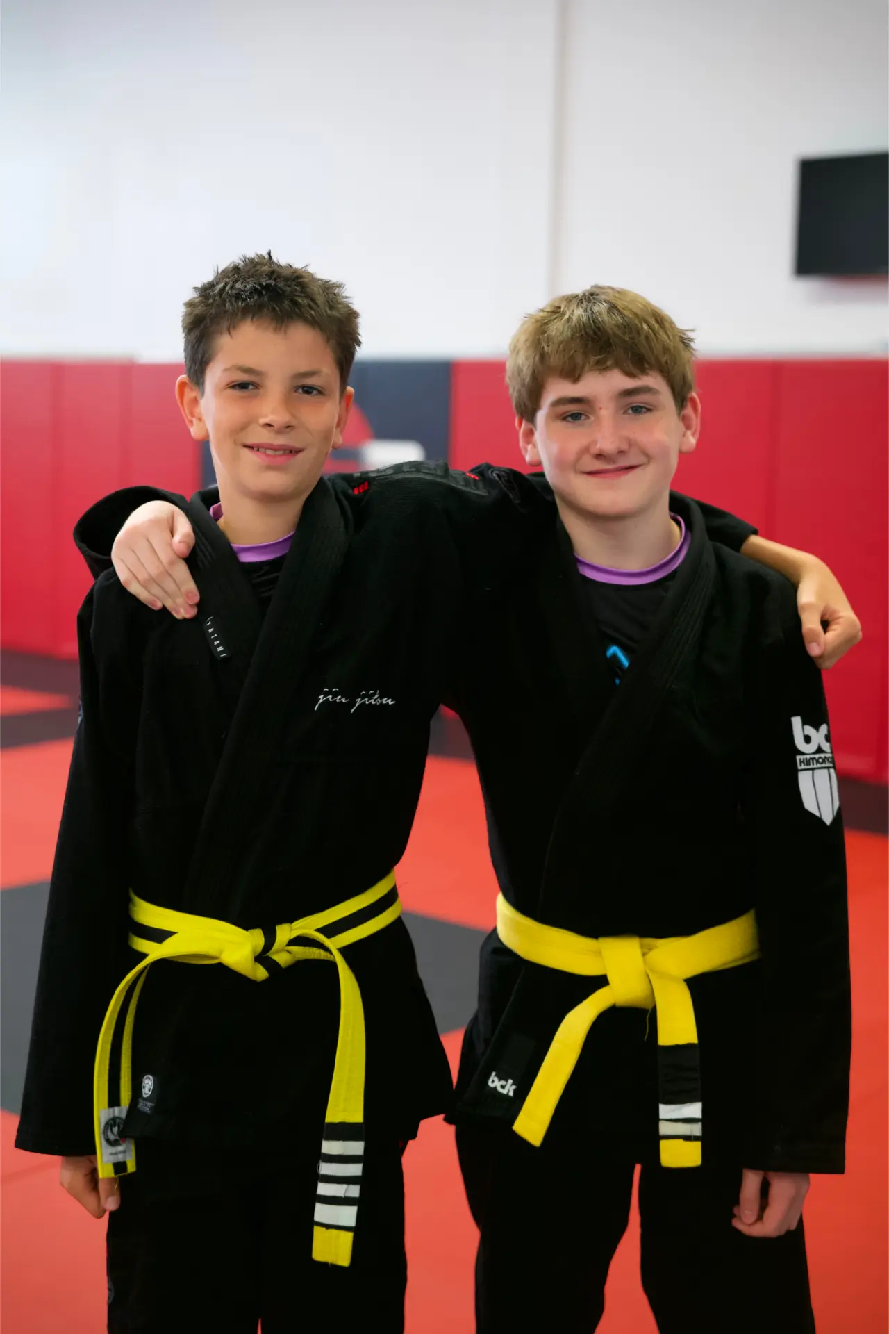 Two teenage Brazilian Jiu Jitsu students in black gis with yellow belts standing arm in arm on the mat at Arashi-Do Red Deer