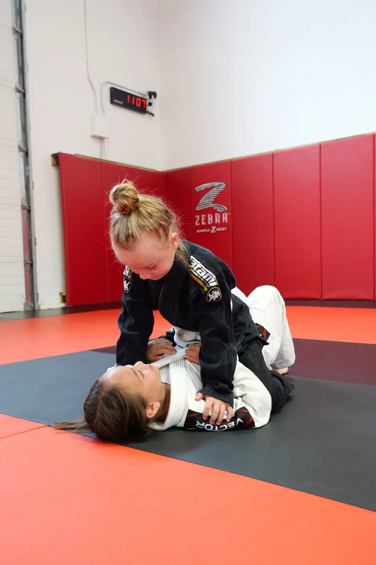 Two young students grappling, one holding top control in a black gi over a partner in a white gi during kids' BJJ class