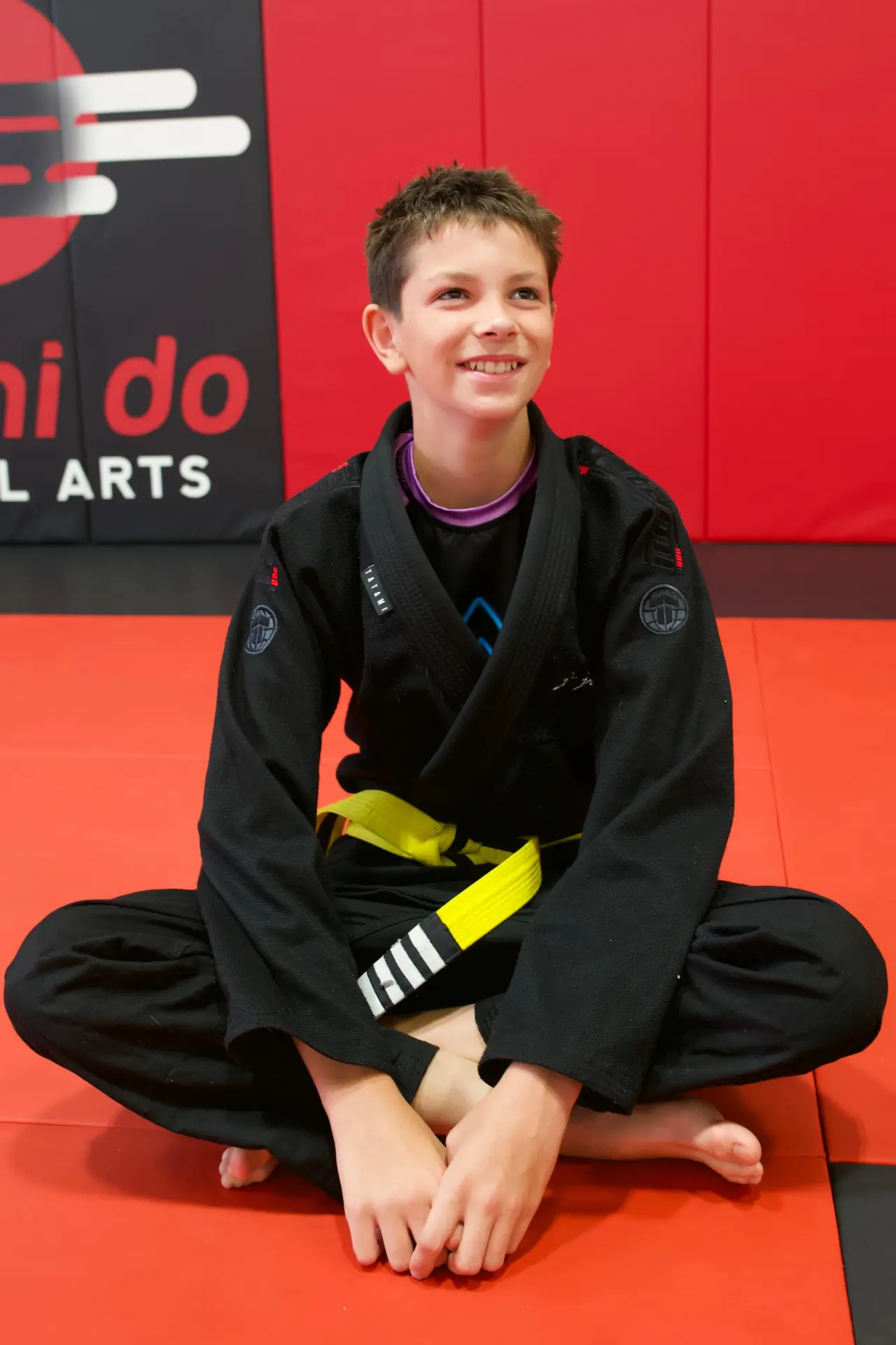 Junior BJJ student in a black gi and yellow belt sitting cross-legged in front of the Arashi-Do Martial Arts wall banner